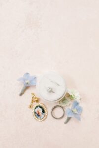 A flat lay of the bride and groom's rings sitting in an ivory velvet ring box surrounded by little flowers and a bouquet charm.