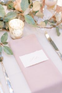 A name card placed on top of a pink napkin at a table with a candle and greenery nearby